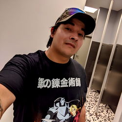 John in a bathroom stall, proudly showing off his t-shirt with anime characters and text on it. He is also tagging his location, perhaps for a social media post. The stall has a simple design, with a white partition visible behind him. 