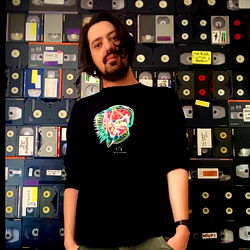 Dan, a model with vibrant hair and colorful clothing standing in front of a collection of VHS tapes.