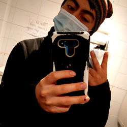 A young adult male from Chile, holding a phone with a selfie of him. He is wearing a face mask and has dark hair.