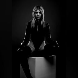 Charley, the fashionable OnlyFans model sitting on a white block in a black outfit against a dark background,