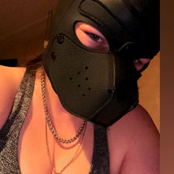 A woman in a black dog mask with her tongue out, wearing a necklace and a sleeveless top,