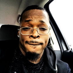 An African American male with glasses and a beard, poking out his tongue in a surprised expression