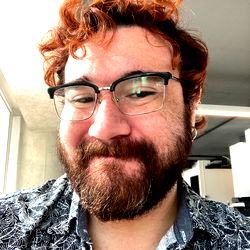 A man with glasses and red hair, bearded, smiling, wearing a shirt and patterned bottoms.