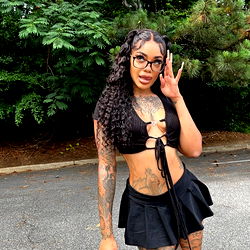 Singer Cream Queen poses in a black and white photograph, showing off her tattoos. She is wearing a denim skirt and has one foot propped up on a car. Her hair is styled in a blonde bob.