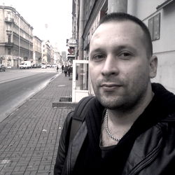 A man posing for a photo on a street corner, near a building with a prominent clock tower.