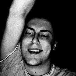 An individual showcasing a visible tattoo in the background, extending their arm and holding a microphone with a smile, seemingly engaged in an audience.