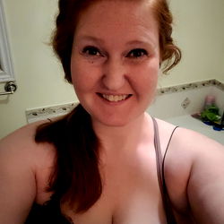 Red-headed woman with dark hair in bra, standing against white wall.