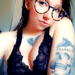 Woman with glasses and tattoos in black lingerie on a bed, posing for OnlyFans