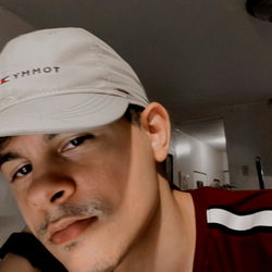 Alekxandre, a young man in his mid-twenties, poses with an intense gaze and slight smile. He's donned in a white cap and sports a modern look.