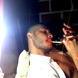 Man in a dark room with a lit cigarette in hand, smoking a joint.