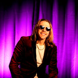 Andrew Rivers in a colorful room with purple lighting and a purple backdrop, wearing sunglasses and a maroon outfit.