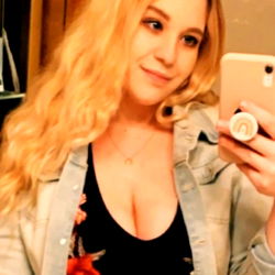 Smiling young woman with long blonde hair, holding a phone and posing for a selfie. Wearing blue jeans, black tank top, and standing in front of a mirror reflection.