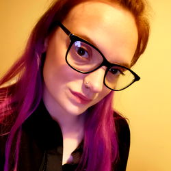 Amber, a vibrant individual with purple hair and glasses, standing in front of a wall