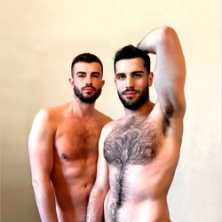 Two men in a studio, one shirtless and the other partially dressed, both with hairy chests