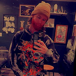 Bearded Man with Long Hair in Hoodie and Hat, Taking Selfie in Tattoo Parlor