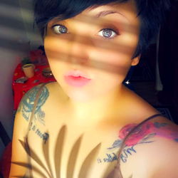 A young adult model with short hair and tattoos, standing in an indoor setting, captured from a side angle.