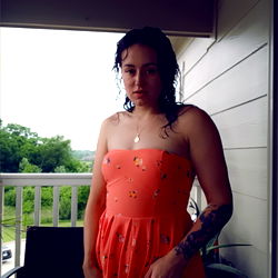 Aphrodite Vixxin poses in an orange dress, standing on a porch