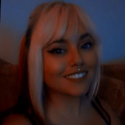 A person with short pink hair and an ear piercing. 1st person standing in front of a mirror with one hand on their hip. They are wearing makeup, including winged eyeliner. The person is smiling and looking directly at the camera.