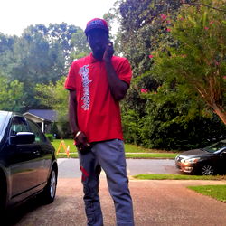 DroSky, an up-and-coming rapper from Tennessee, poses on the sidewalk with a red shirt and black pants. He holds a phone to his ear and is looking directly at the camera. His surroundings include trees, cars, and buildings in the background.