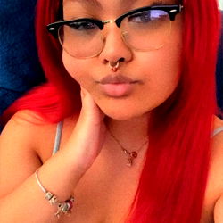 A young woman with vibrant red hair and glasses, wearing a sleeveless top, poses confidently for the camera. She's sitting on a couch.
