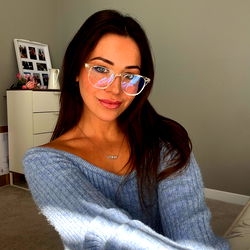 A young woman in blue sweater, wearing glasses and blue nail polish, poses with one hand on her hip against a light backdrop.