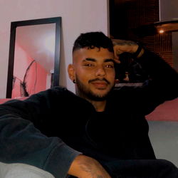 Alejandro Delgado, a young man with short hair, is sitting on a sofa and smiling. He's wearing a black jacket and has his arm resting on the armrest of the couch. A mirror hangs behind him. The room appears to be well-lit and inviting.