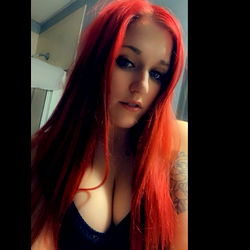 Red-haired woman with tattoos and a piercing, posing in a black bra and red lipstick