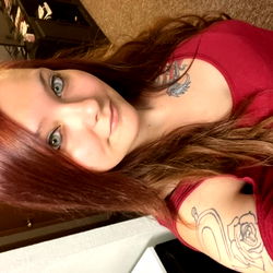 Ally, a beautiful red-haired woman with tattoos and piercings. She is seen in a vibrant red top.