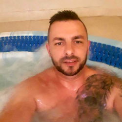 A man enjoying a relaxing moment in a hot tub, with a tattoo visible on his left arm. He appears to be in good spirits and is smiling towards the camera.