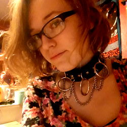 A young woman in glasses with a choker necklace and rings around her neck posing for the camera.