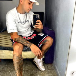Young man with tattoos, holding a smartphone and leaning against a wall in a room, taking a selfie. He is wearing a white cap, a t-shirt, shorts, and sneakers. In the background, there's a bed and a mirror reflecting the room.