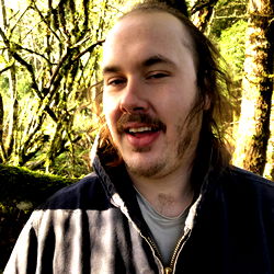 Bearded man in a forest setting, wearing a dark jacket and glasses.