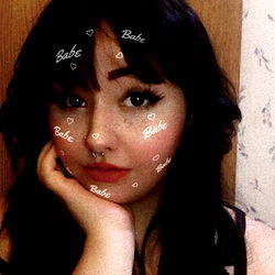 A young woman with a tattoo, seated indoors in front of a wallpaper-covered wall. She has dark hair and is looking directly at the camera.