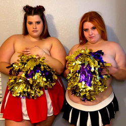Two girls, one of whom appears to be in a cheerleading uniform, standing together in a room with walls featuring a wallpaper pattern. They are both smiling and seem to be in good spirits.