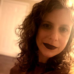 Emire, a young woman with dark hair, is striking an edgy pose with her tongue out. Her style is unique and fashionable, characterized by dark makeup around her eyes. She sports a chic bob hairstyle and has a tattoo on her right wrist.