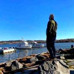 An individual at a waterside with boats and dock in the background, standing on rocks and looking into the distance.