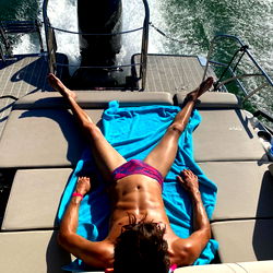 A shirtless man on a boat, lying on his stomach with his head turned to one side. He is wearing blue shorts and pink swim trunks.