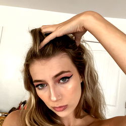 Aly, a young adult woman, poses confidently with one hand on her head and the other holding a hair accessory. She is wearing makeup and has blonde hair. Her attire consists of a casual t-shirt.