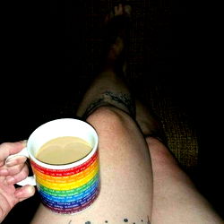 Kitten, a tattooed individual with pride flag on her thighs enjoying a cup of coffee in a cozy setting.