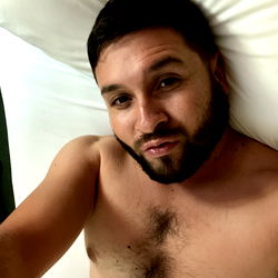 Sam, a social media influencer lying on his stomach in bed with tattoos and a beard, showcasing his fit physique.