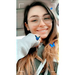Anna, a young woman with glasses and butterflies in her hair, smiles at the camera from inside a car. Age: 25, Gender: Female
