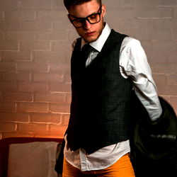 A dapper young man in a stylish outfit and glasses, standing confidently in a room with a brick wall.