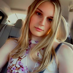 Lucy, a young woman with blonde hair, captured in a candid moment inside a car, wearing a floral top and accessorized with a bracelet.