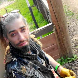 WittyMire, a bearded man in a bandana and work clothes leaning on a fence,