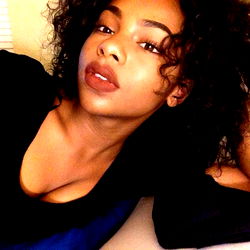 Lexii, a beautiful black woman with natural hair and lipstick, sitting and looking at the camera.