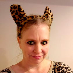 A playful woman wearing a leopard cat ear headband, striking a fun pose in front of a white wall, with her hands on her hips.