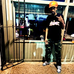 TheBigDill posing outside a storefront in camouflage pants and a black t-shirt, with a playful finger-pointing gesture