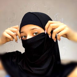 Woman in a hijab holding face mask with hands, wearing black dress and nail polish