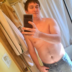 A young man poses shirtless for a mirror selfie in his bathroom.