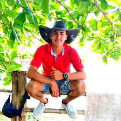 A young man posing outdoors with a casual and relaxed demeanor, wearing a cowboy hat and a red shirt.